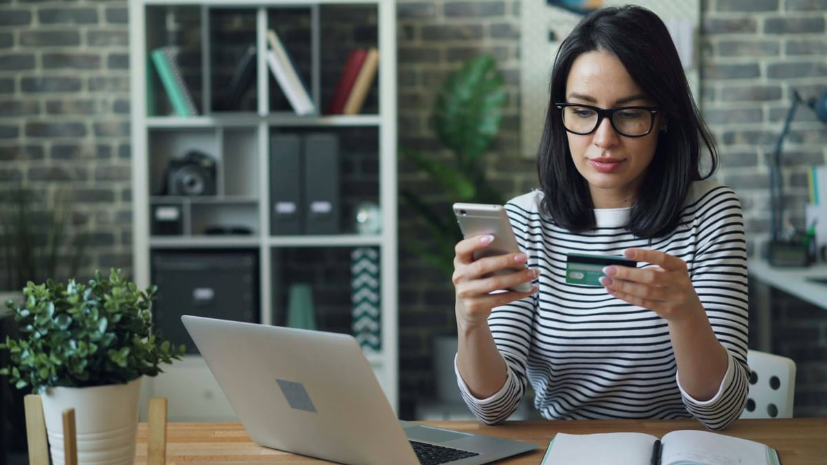 Woman using a smartphone with a credit card while working at a laptop